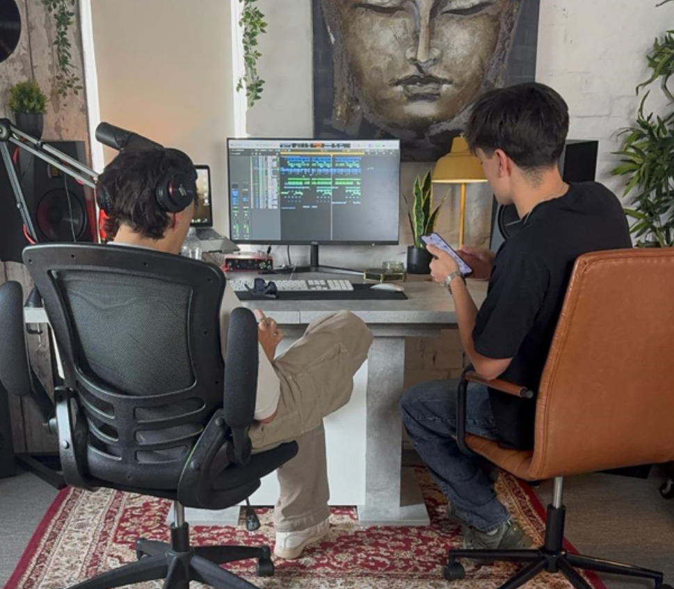 Two people working together at the desk, seen from behind, with a Buddha painting and hanging plants on the wall above the monitors.