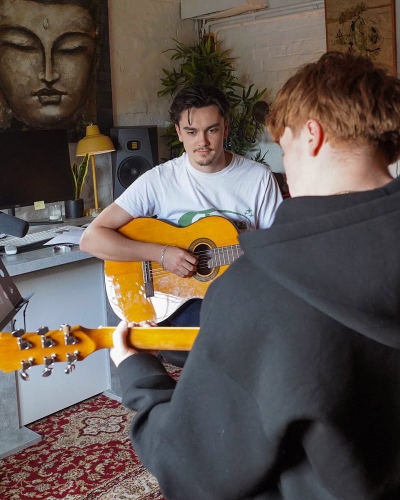 Josh English in the studio playing an acoustic guitar, facing a student who is following along on their own guitar.